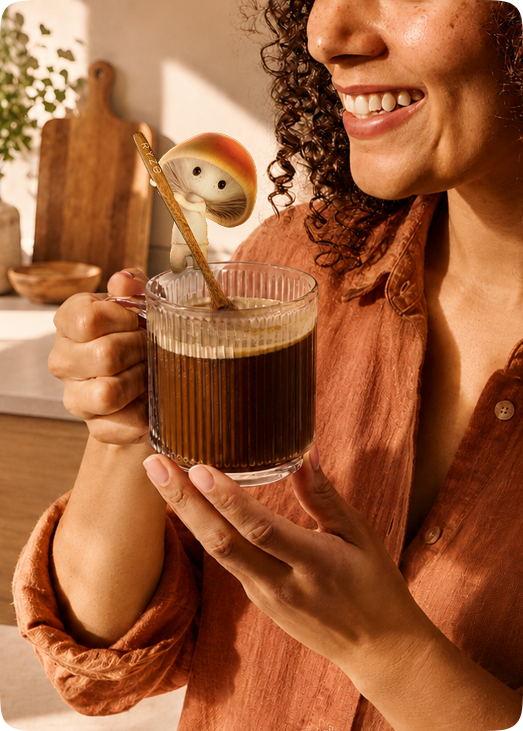 A smiling woman in a rust-colored shirt holds a clear ribbed glass mug filled with dark coffee. A whimsical mushroom-shaped spoon rests in the mug, and the background shows a warm, softly lit kitchen scene with wooden cutting boards and a plant.