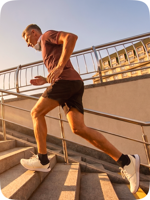 A man in a maroon t-shirt, black shorts, and white sneakers running up outdoor concrete stairs at sunset.