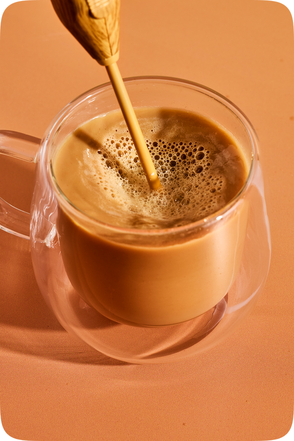 A clear glass mug filled with a frothy light brown beverage, set against a warm peach background with a wooden spoon resting inside the cup.