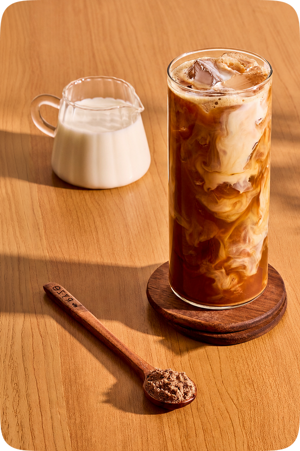 A tall glass of iced coffee with milk swirls and ice, served on a round wooden coaster on a warm wooden table. A small glass pitcher of milk sits behind the glass, and a wooden spoon with brown sugar rests in the foreground.