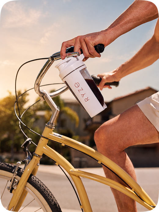 A close-up lifestyle shot of a person riding a yellow bicycle outdoors, holding a white shaker bottle with the brand name 'RYZE' printed vertically on the side.