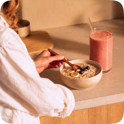A person in a white robe is shown from the side, holding a wooden spoon over a bowl of oatmeal with banana slices and blueberries. A glass of pink smoothie sits on the counter beside the bowl in a warm, softly lit kitchen setting.