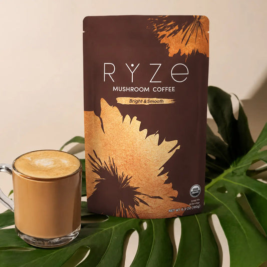 A pouch of RYZE Mushroom Coffee stands upright on green monstera leaves next to a glass mug filled with a latte, against a warm beige background.