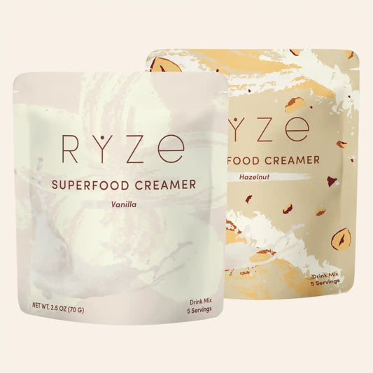 Two stand-up pouches of RYZE Food Creamer shown on a plain light beige background. The front pouch is Vanilla and is mostly white with soft pale gray marbling; the rear pouch is Hazelnut and has a warm beige/cream base with tan and brown nut-piece accents.