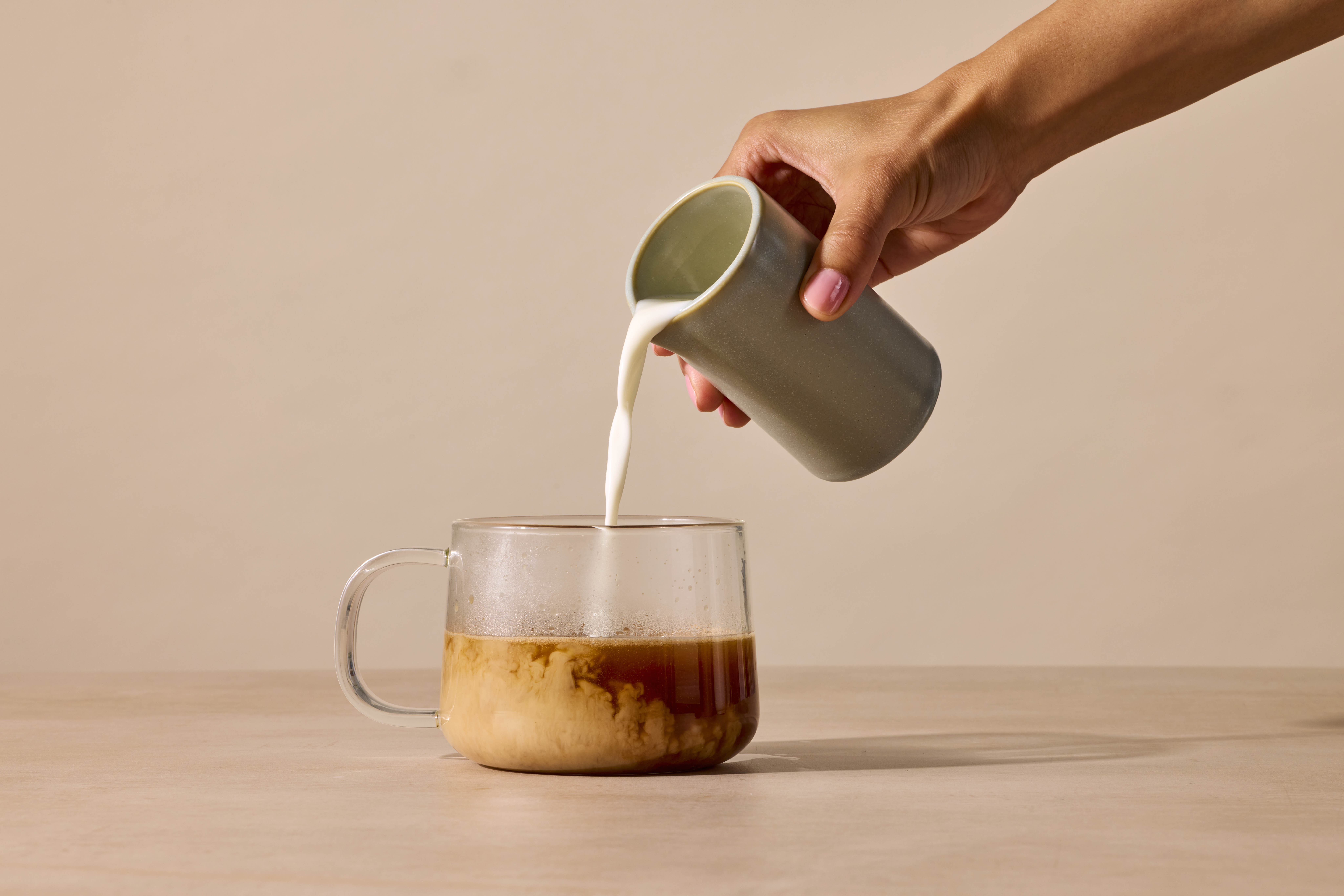 Hand pouring milk into a glass mug of coffee.