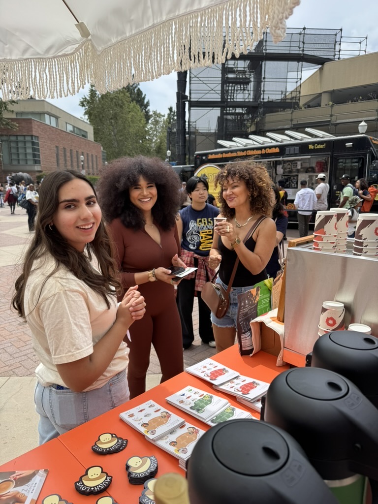 Three women smiling at an outdoor market stall with brochures and coffee dispensers.